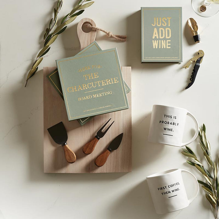 Lifestyle photograph of charcuterie and wine gift accessories on a marble surface featuring sage green boxes with gold foil typography, wooden-handled cheese knives, and white ceramic mugs with humorous wine quotes beside olive branches