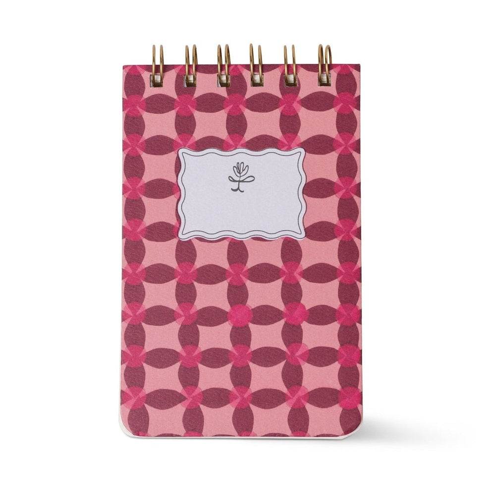 Spiral-bound paper notebook featuring a repeating pink and maroon geometric floral pattern, a central scalloped white label with a minimalist flower icon, and a gold-toned metal spiral binding against a plain white background
