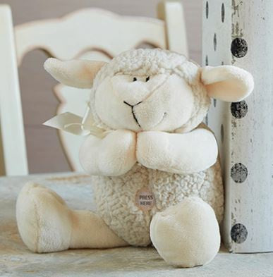 Photograph of a Hallmark plush praying lamb sitting on a tabletop in front of a white chair featuring textured cream-colored fleece, a small white ear ribbon, and a circular Press Here button on its chest