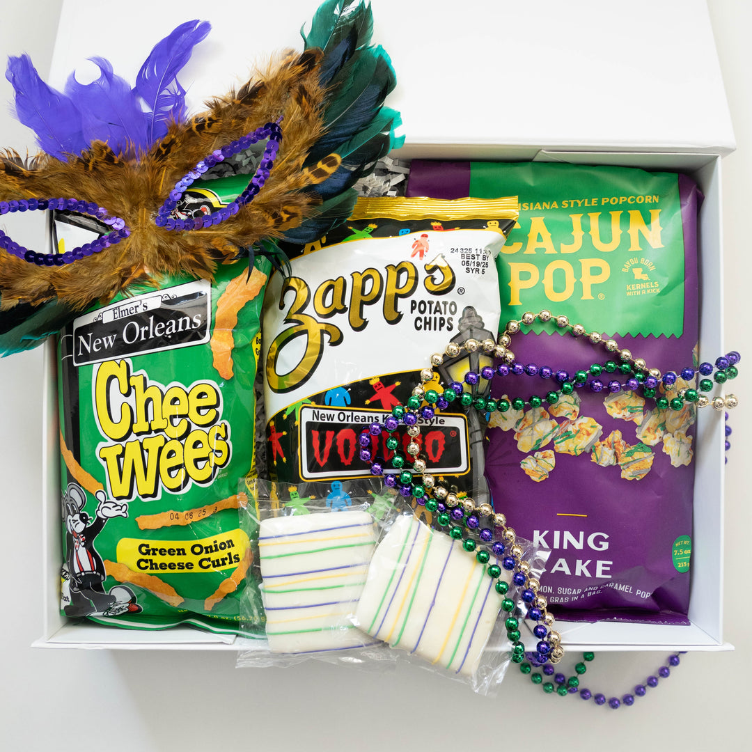 Photograph of a New Orleans Mardi Gras gift box featuring Zapp's Voodoo potato chips and Elmer's Chee Wees alongside a vibrant purple feathered mask and colorful metallic beads
