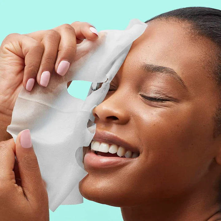 Studio photograph of a smiling Black woman peeling a damp white sheet mask from her face against a vibrant teal background, showcasing her pale pink manicured nails and radiant skin