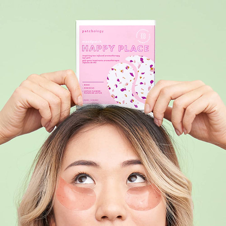 Studio photograph for Patchology featuring a woman with ombre blonde hair wearing translucent pink eye gels and holding a Patchology Happy Place moodpatch box with colorful floral patterns against a mint green background