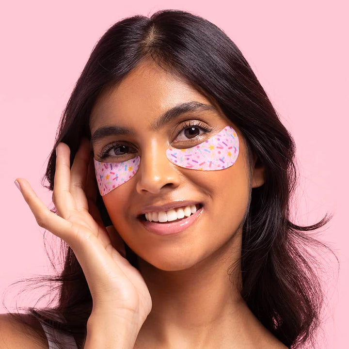 Portrait photograph of a smiling young woman with dark hair against a vibrant pink background featuring colorful sprinkle-patterned under-eye patches and a hand resting on her cheek