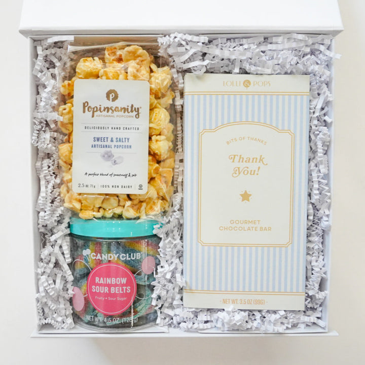 Digital photograph of a white gourmet snack gift box featuring golden-brown Popinsanity popcorn, colorful Candy Club rainbow sour belts, and a blue-striped Lolli &amp; Pops gourmet chocolate bar nestled in white shredded paper filler