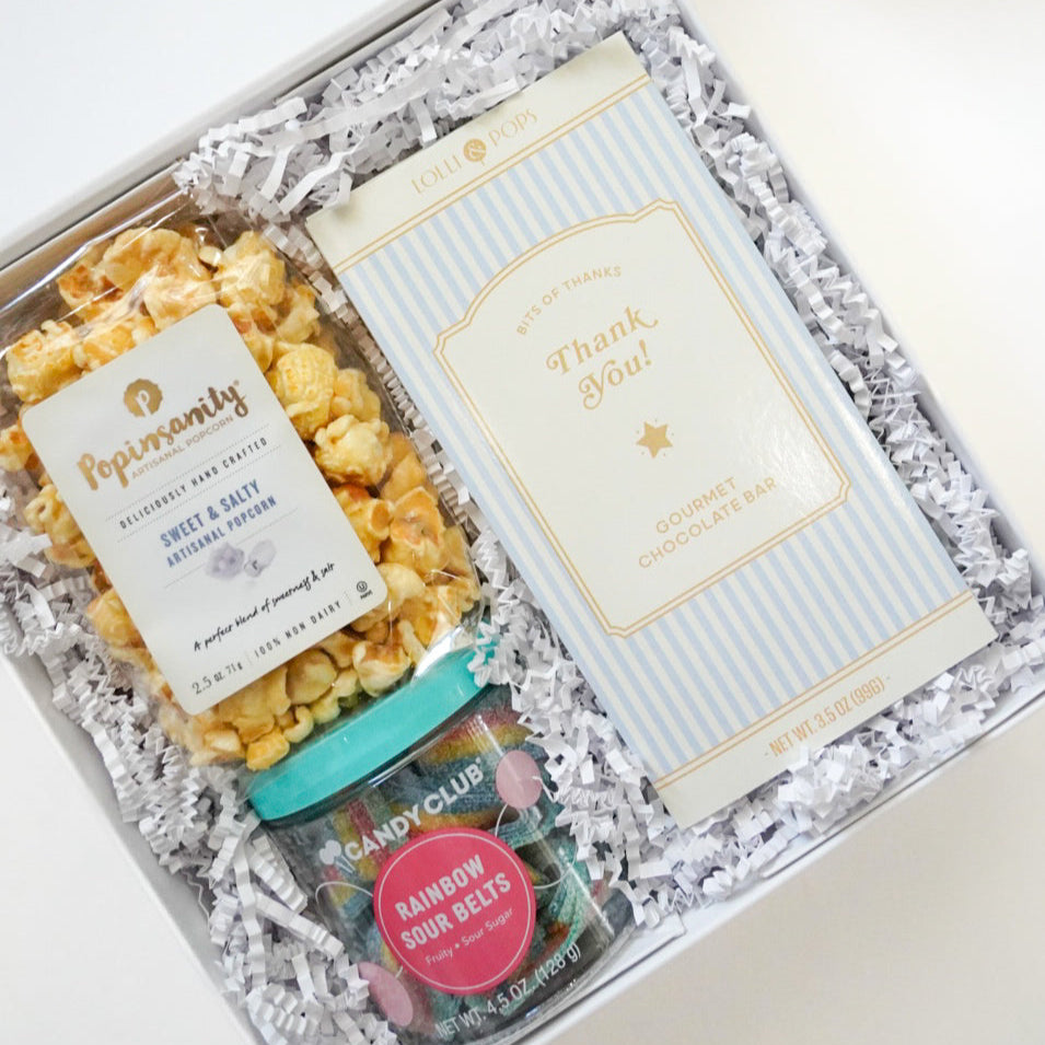 Photograph of a gourmet thank you gift box containing a bag of Popinsanity sweet and salty popcorn, a blue striped Lolli and Pops chocolate bar, and a jar of rainbow sour belts nestled in white crinkle paper