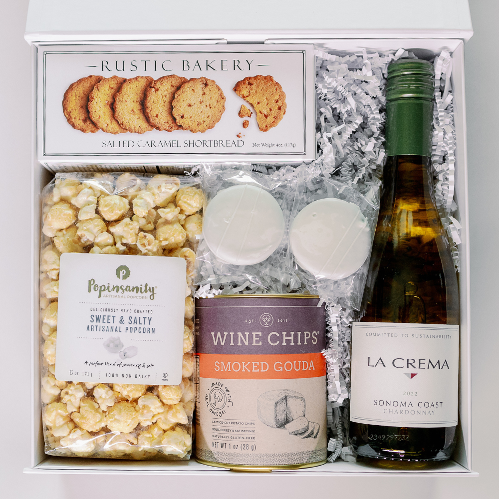 Photograph of a curated gourmet gift box featuring a bottle of La Crema Sonoma Coast Chardonnay, a bag of golden Popinsanity popcorn, and an orange-labeled canister of smoked gouda Wine Chips nestled in white crinkle paper