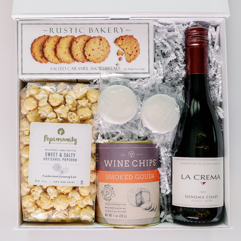 Digital photograph of a curated gourmet gift box containing a bottle of La Crema Pinot Noir, Rustic Bakery salted caramel shortbread, and Popinsanity artisanal popcorn nestled in white crinkle paper alongside a tin of Smoked Gouda wine chips and two white chocolate treats