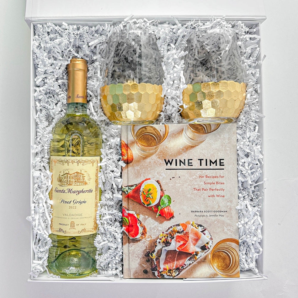 Digital photograph of a premium wine gift box featuring a bottle of Santa Margherita Pinot Grigio, a Wine Time cookbook with colorful food imagery, and two stemless glasses with gold honeycomb bases nestled in white shredded paper