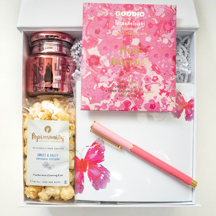 Digital photograph of a curated pink gift box featuring Goodio x Ivana Helsinki floral chocolate, a metallic rose-gold jar candle, Popinsanity popcorn, and a gold-trimmed pink pen resting on butterfly-adorned cards