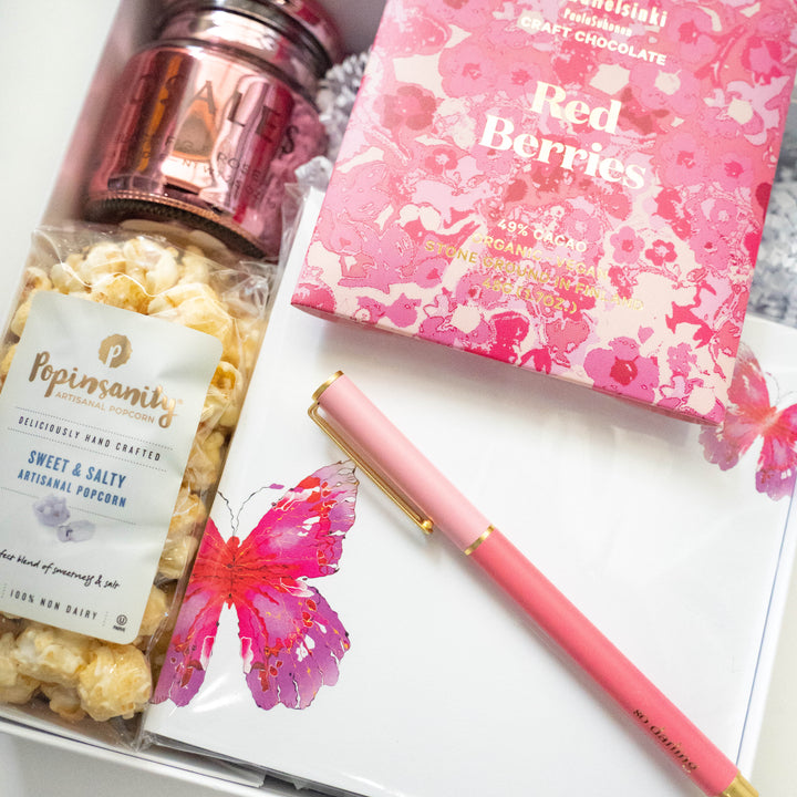 Photograph of a curated gift box showcasing Popinsanity popcorn, Red Berries craft chocolate, a rose gold jar, and a white card with a vibrant watercolor butterfly and a pink-and-gold pen