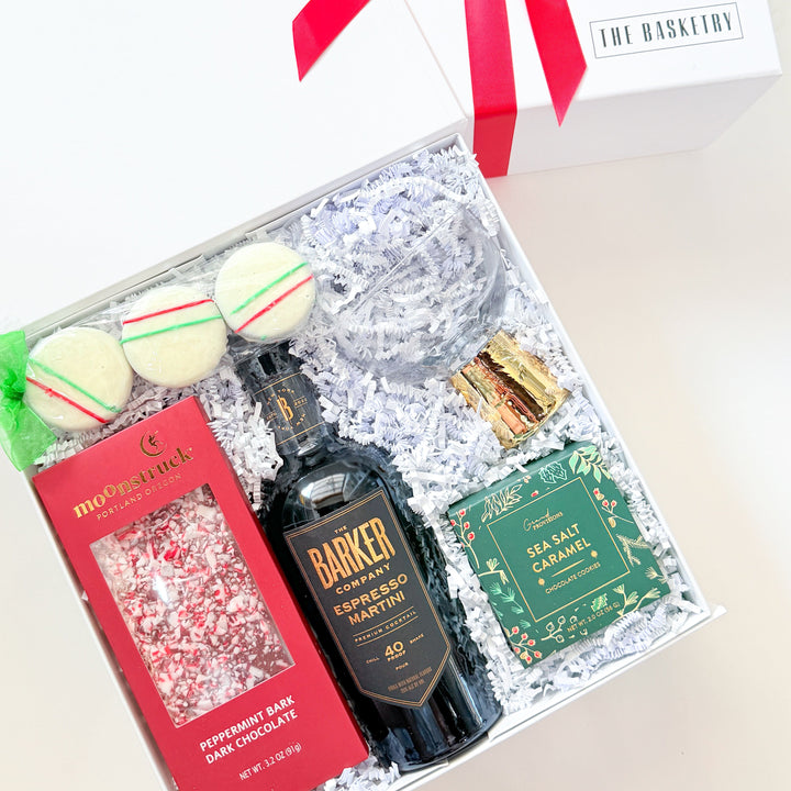 An open white gift box tied with a red ribbon, filled with assorted gift items on white crinkle paper. Visible contents include a dark bottle labeled Barkey&