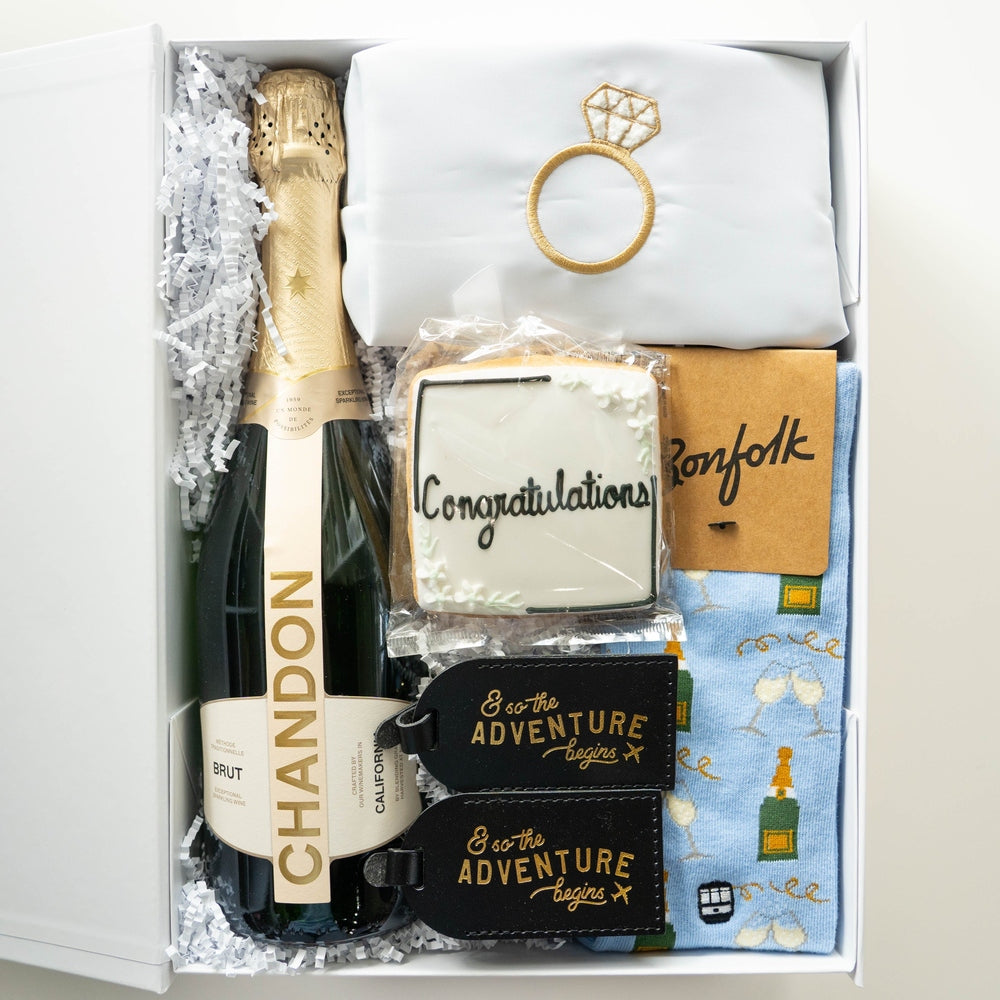 Photograph of an engagement celebration gift box featuring a bottle of Chandon Brut, a white fabric with a gold-embroidered diamond ring, a square iced Congratulations cookie, and black leather luggage tags with gold script