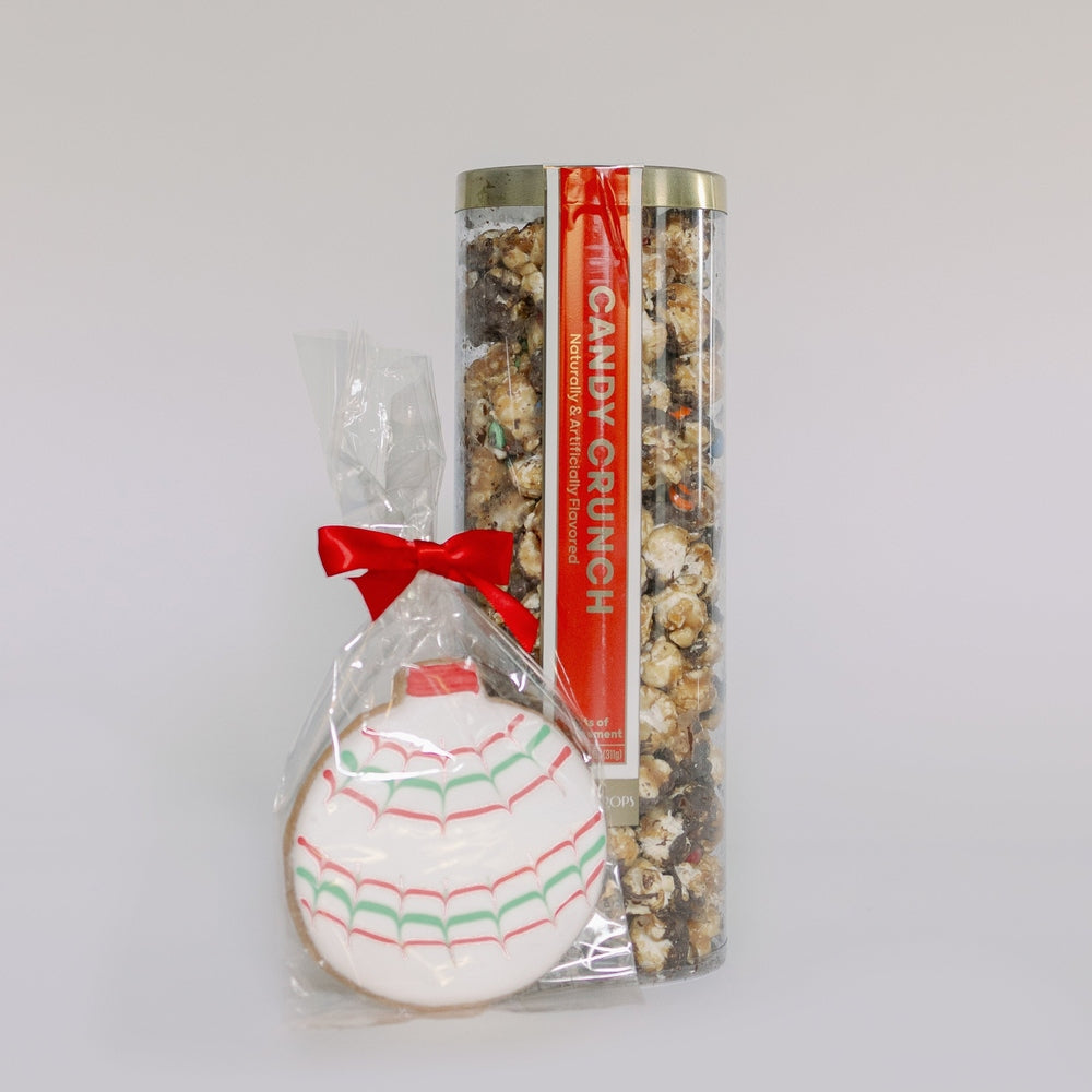 Commercial product photograph of a tall Candy Crunch snack canister and an ornament-shaped sugar cookie on a white background, featuring a bright red ribbon bow and white icing with red and green scalloped patterns