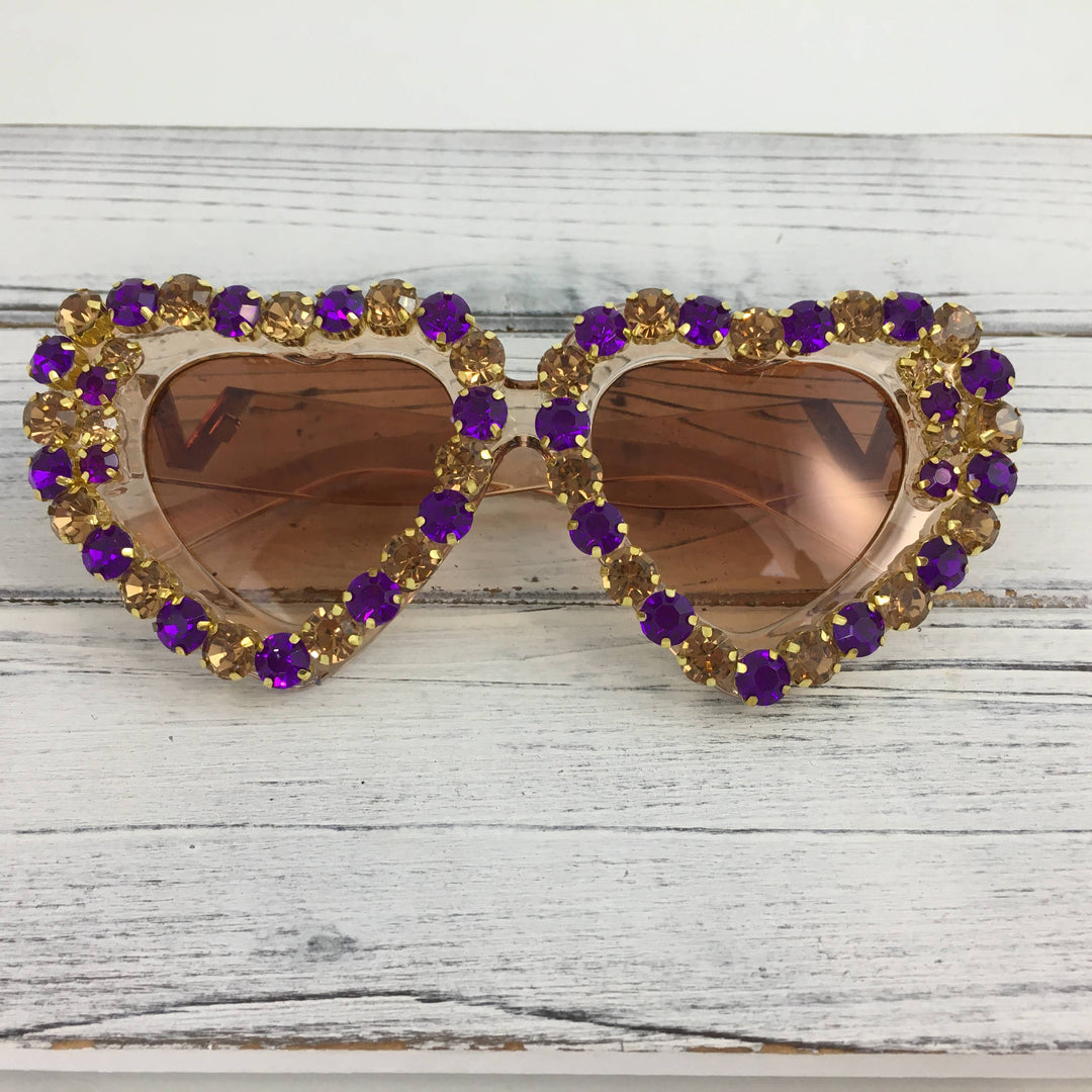 Hand-embellished heart-shaped sunglasses resting on a white-washed wooden surface featuring a decorative border of alternating purple and amber rhinestones and warm brown-tinted lenses