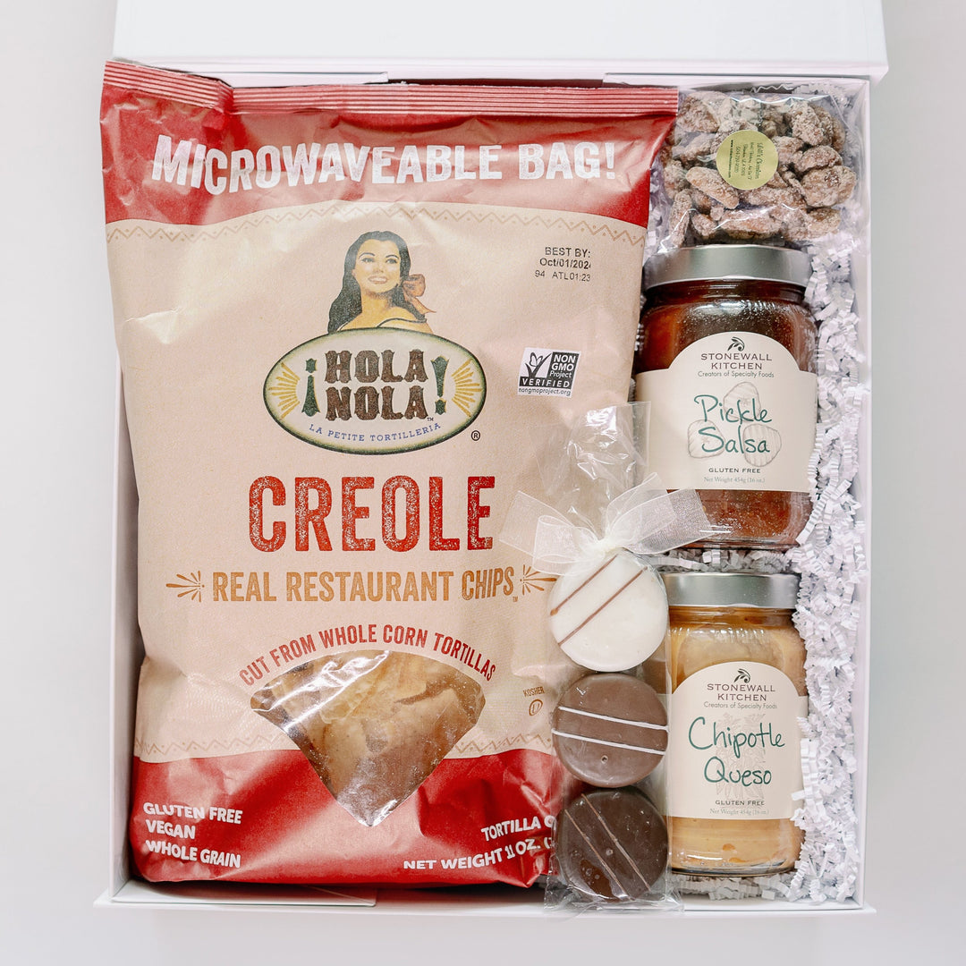 Photograph of a gourmet snack gift box containing a large red and tan bag of Hola Nola Creole tortilla chips, jars of Stonewall Kitchen salsa and queso, and three striped chocolate treats tied with a white organza bow nestled in white crinkle-cut paper filler