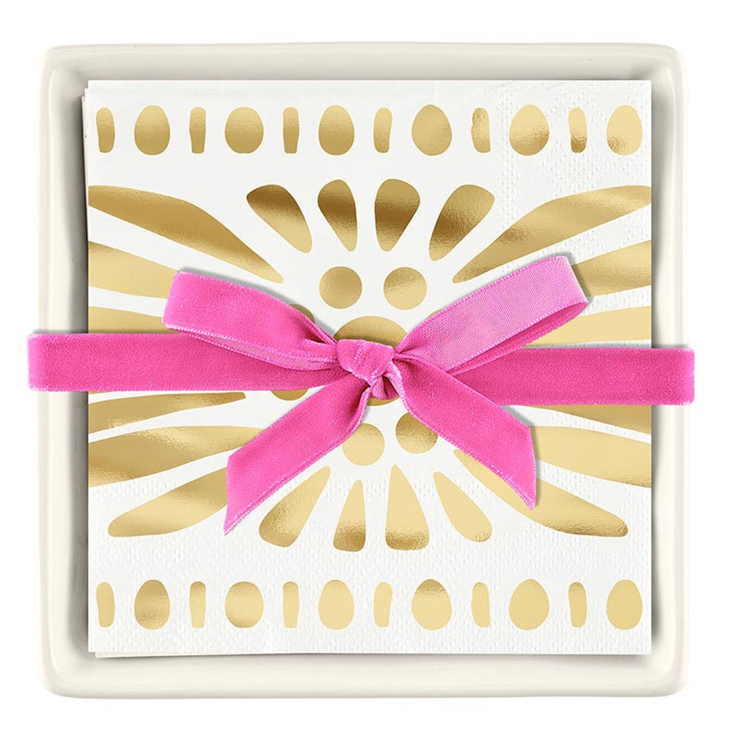 Decorative paper cocktail napkins featuring a metallic gold sunburst pattern are displayed in a square white ceramic tray and tied with a vibrant pink velvet ribbon bow
