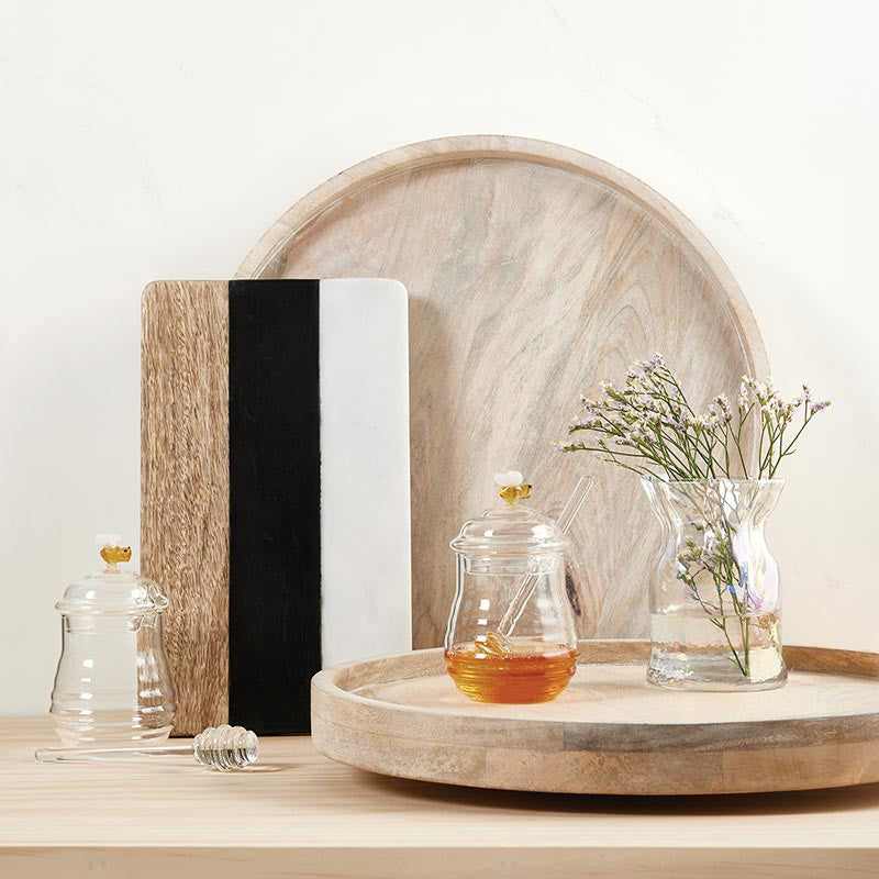 Professional lifestyle photograph of a kitchenware set on a light wood surface featuring round mango wood trays and a vertical tricolor marble board alongside glass honey pots with tiny yellow bee figurines and a small bouquet of purple flowers in an iridescent vase