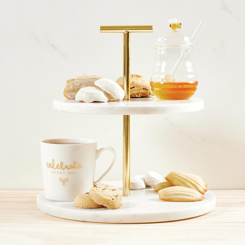 Professional product photography of a two-tiered white marble and gold-tone serving stand featuring a glass honey pot with a yellow bee lid, a white mug with gold celebrate every day script, and classic French madeleines on a light wood tabletop