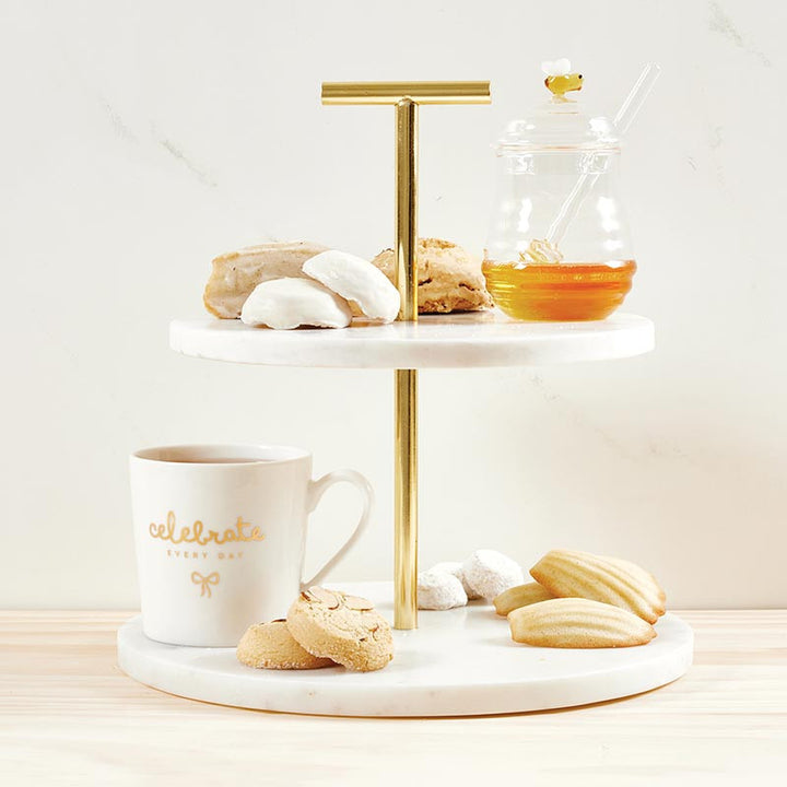Professional product photography of a two-tiered white marble and gold-tone serving stand featuring a glass honey pot with a yellow bee lid, a white mug with gold celebrate every day script, and classic French madeleines on a light wood tabletop