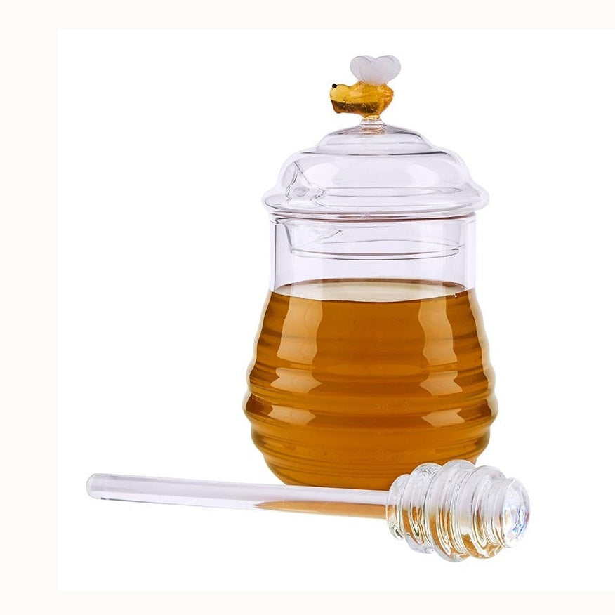 Glass honey pot and dipper on a white background featuring a ribbed beehive-shaped jar filled with golden honey and a small glass bee figurine on the lid