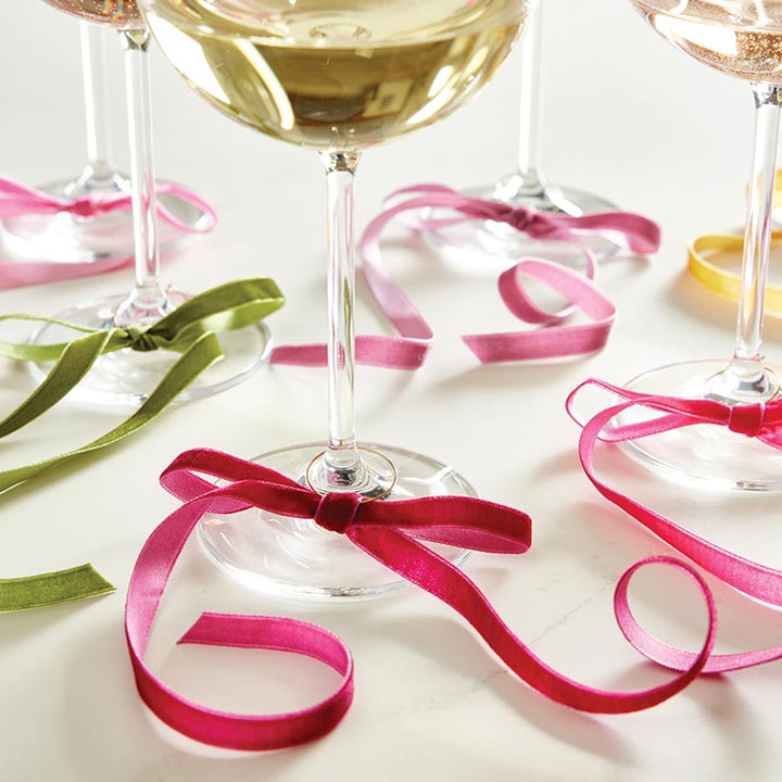 Digital photograph of elegant champagne glasses on a white surface featuring vibrant fuchsia, olive green, and light pink velvet ribbons tied in bows around the stems