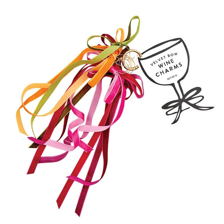 Product photography of Joanna Buchanan set of six velvet bow wine charms featuring vibrant magenta, olive green, and burnt orange ribbons attached to thin gold-tone rings beside a white wine glass-shaped display card