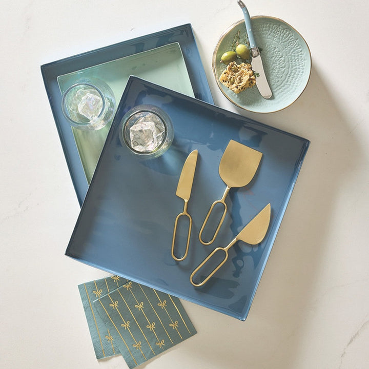 Lifestyle photograph of a modern tableware arrangement on a white marble surface featuring square enameled trays in blue and mint green, matte gold cheese knives with oval loop handles, and teal napkins printed with gold bow patterns