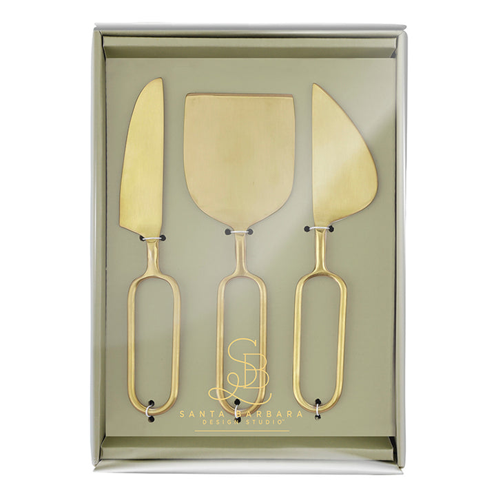 Brass-finished stainless steel cheese tool set by Santa Barbara Design Studio featuring three distinct serving knives with minimalist elongated oval loop handles displayed inside a sage-green gift box with a gold-foil logo