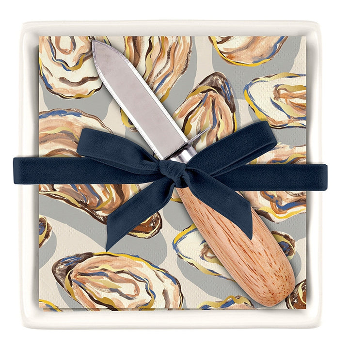 Painterly illustration by Caspari of an oyster-patterned cocktail napkin and wooden-handled knife gift set featuring a navy blue ribbon bow and gold-flecked shell motifs inside a white ceramic dish