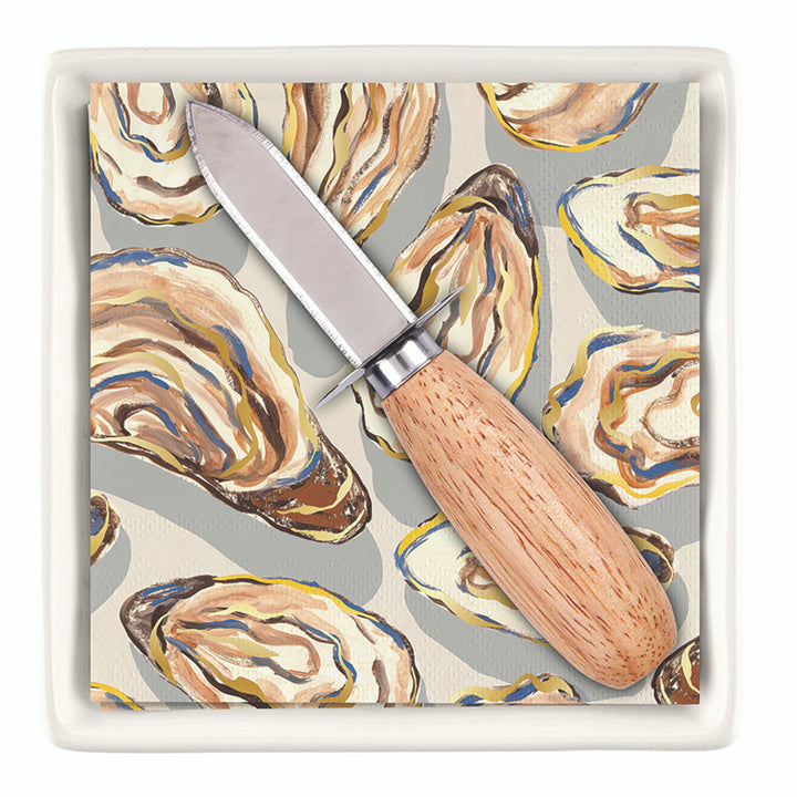 Digital product photograph by Michel Design Works of a stainless steel oyster knife with a light wood handle resting on paper napkins featuring painterly blue and yellow oyster shell illustrations inside a white square ceramic tray