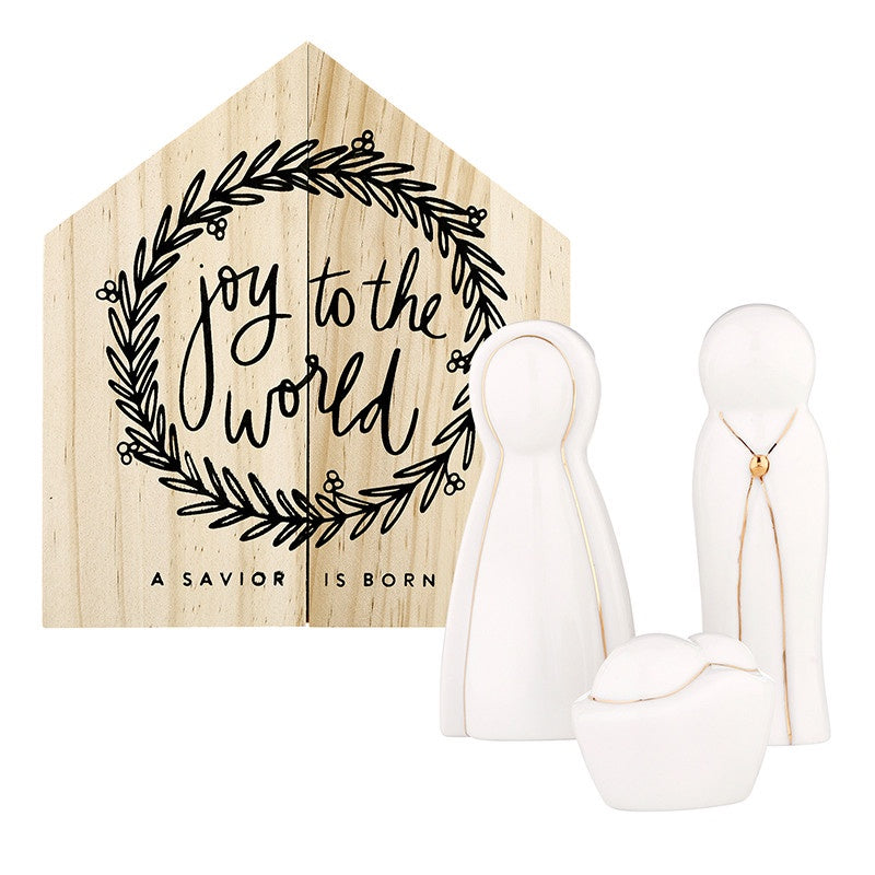 Ceramic and wood Mud Pie nativity set depicting the Holy Family in front of a peaked wooden creche backdrop featuring a black laurel wreath, faceless white figurines with gold wire accents, and hand-lettered joy to the world calligraphy