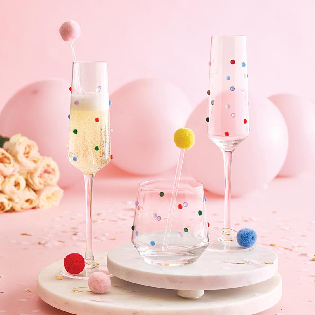 Studio photograph of champagne flutes and a lowball glass adorned with vibrant multi-colored dots set on white marble platters against a festive backdrop of pink balloons, soft roses, and whimsical pom-pom drink stirrers