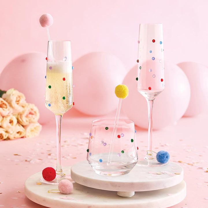 Studio photograph of champagne flutes and a lowball glass adorned with vibrant multi-colored dots set on white marble platters against a festive backdrop of pink balloons, soft roses, and whimsical pom-pom drink stirrers