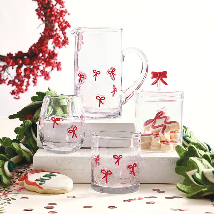 Hand-painted glassware set featuring a clear pitcher and tumblers adorned with delicate red bows on white holiday pedestals surrounded by a vibrant red berry wreath and a sugar cookie iced with the word JOY