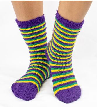 Photograph of plush Mardi Gras themed socks worn on feet showcasing horizontal purple green and yellow stripes with fuzzy solid purple toes and ankle cuffs against a white studio backdrop