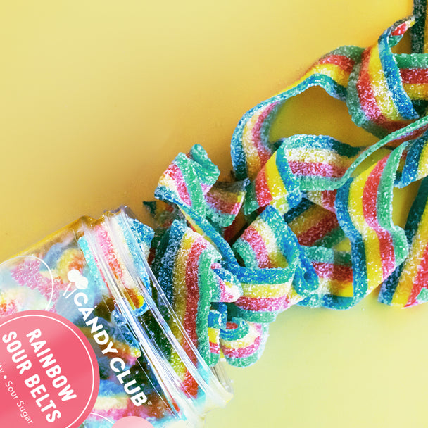 Commercial photograph by Candy Club of Rainbow Sour Belts spilling from a clear jar onto a bright yellow surface featuring sugar-dusted ribbons with vibrant multicolored stripes and a circular pink brand label