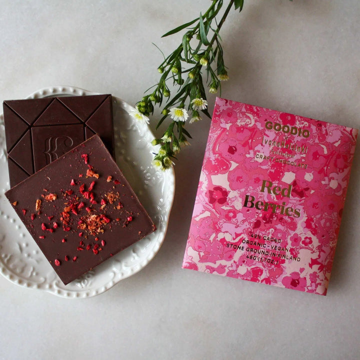 Photograph of Goodio and Ivana Helsinki craft chocolate featuring a dark chocolate bar topped with dried red berry bits on a white scalloped plate next to pink floral packaging and a sprig of white wildflowers on a light stone surface