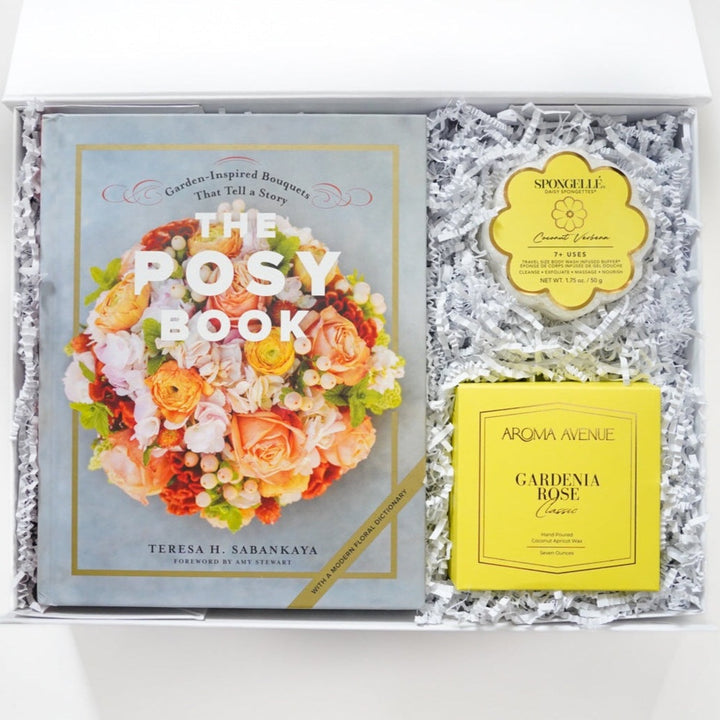 Photograph of a curated floral gift box featuring The Posy Book by Teresa H. Sabankaya, a yellow daisy-shaped Spongellé buffer, and a yellow Aroma Avenue Gardenia Rose candle box nestled in white shredded crinkle paper