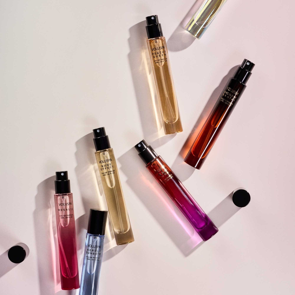 Digital product photography by Voluspa featuring an array of slender glass perfume vials scattered across a light beige surface including a vibrant magenta bottle and a cornflower blue Neroli Sunbath spray casting sharp directional shadows