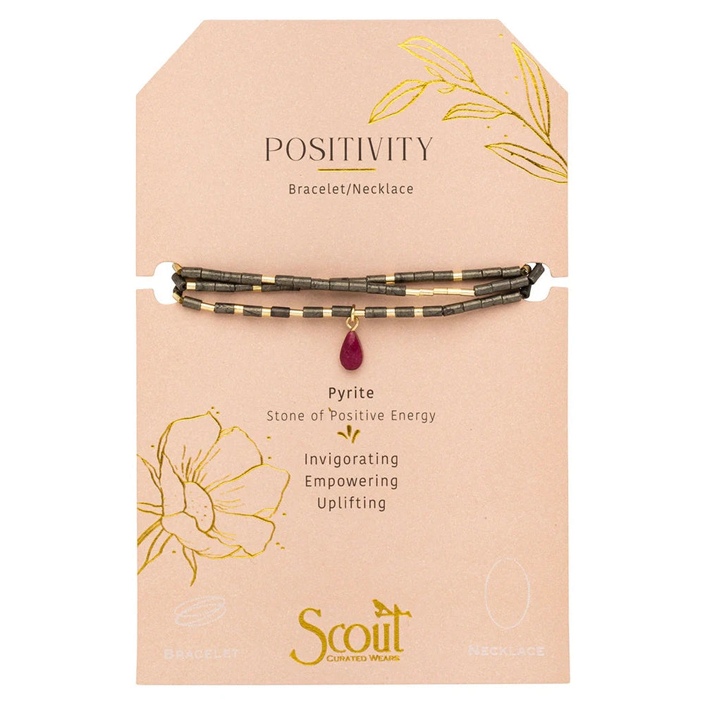 Scout Curated Wears jewelry features a multi-strand pyrite bead bracelet-necklace centered on a peach Positivity display card featuring a teardrop-shaped magenta stone pendant and delicate gold foil botanical illustrations