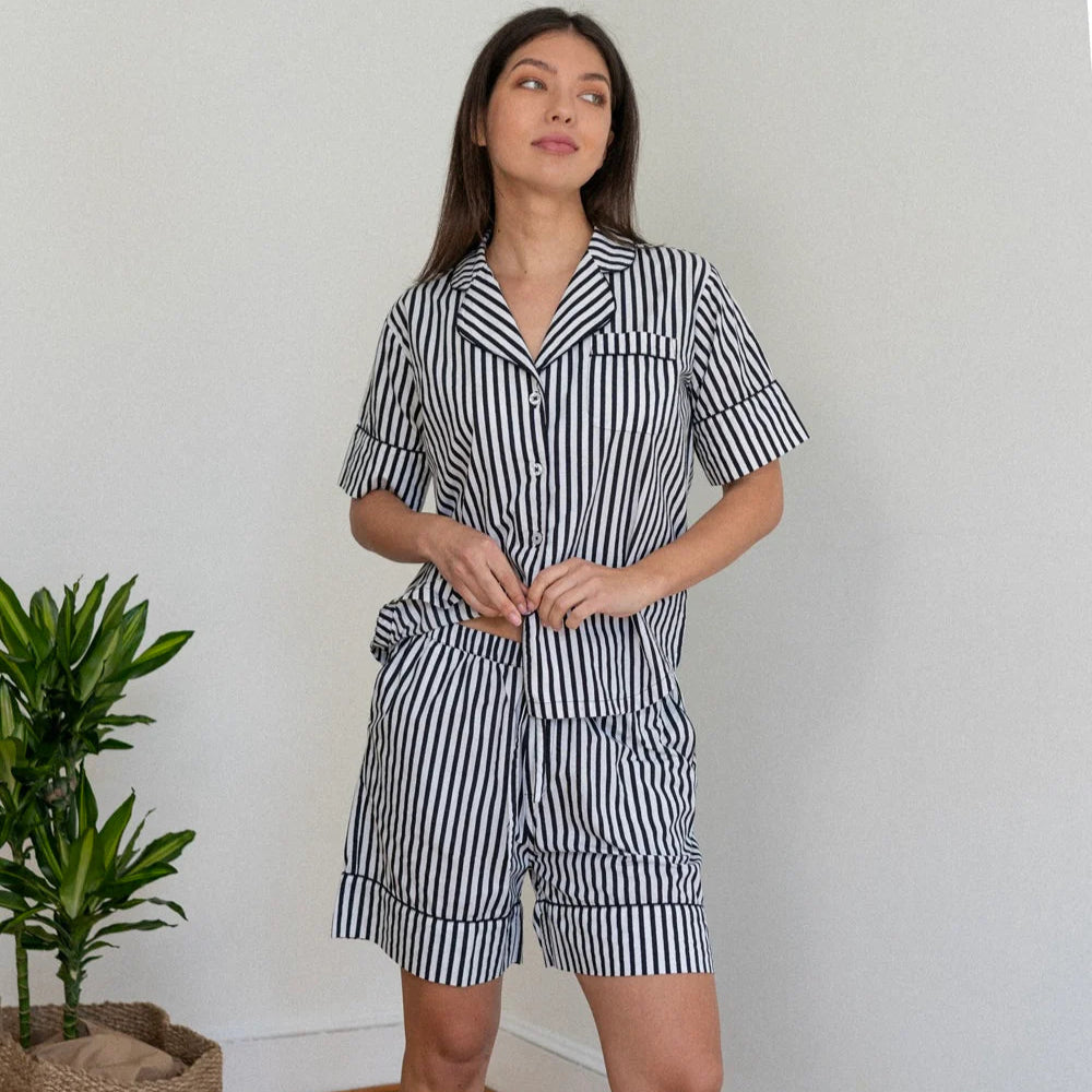 Photograph of a woman wearing a navy and white vertically striped short-sleeve pajama set in a minimalist room featuring a notched collar and a green leafy plant in a woven basket