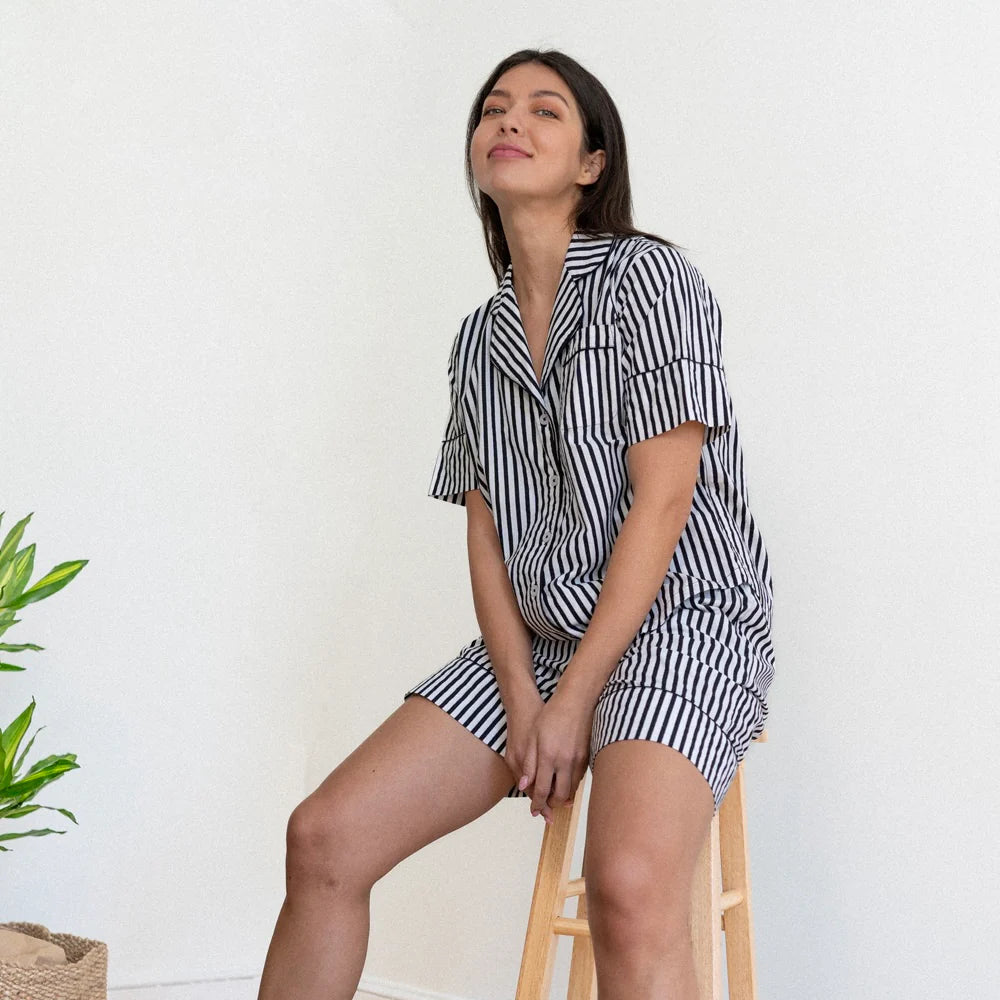 Digital photograph of a woman sitting on a light-colored wooden stool in a white studio wearing a black and white vertical striped pajama set with a notched lapel and a vibrant green plant in the corner