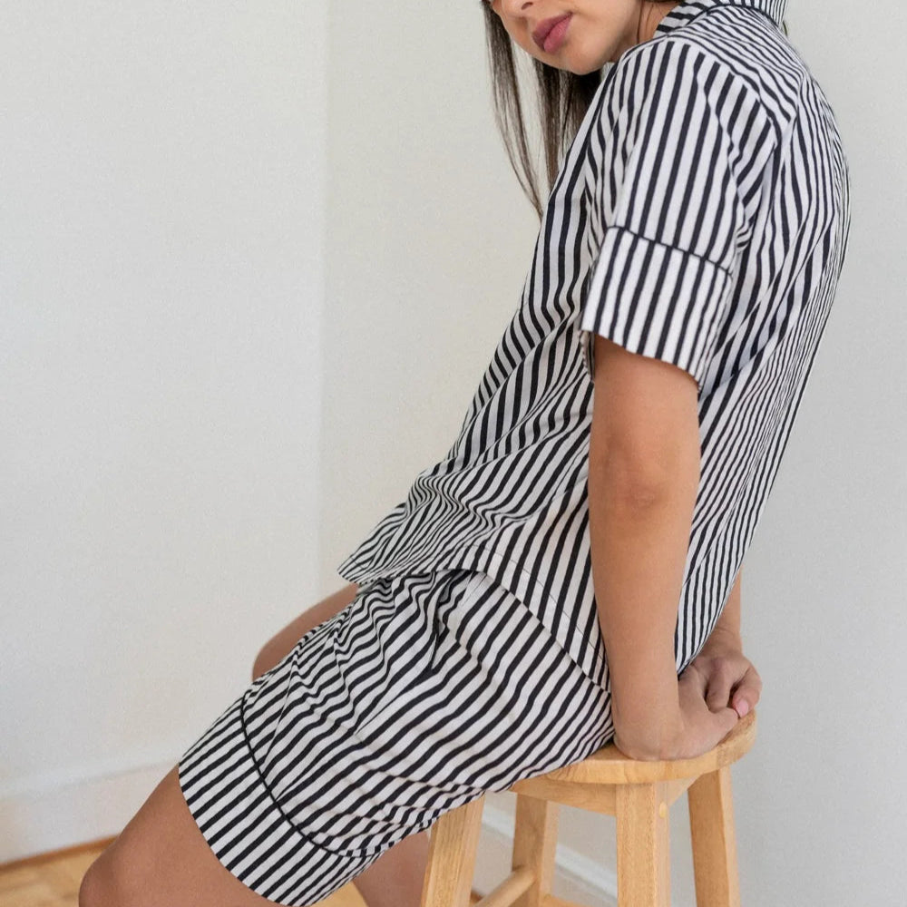 Photograph of a woman wearing a black and white vertical striped pajama set while sitting on a light wood stool in a minimalist white corner featuring cuffed short sleeves and a high-contrast pattern