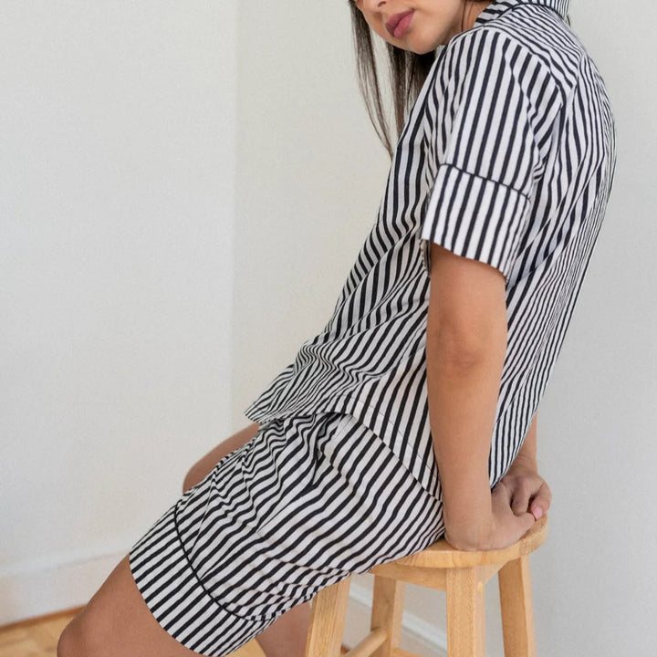 Photograph of a woman wearing a black and white vertical striped pajama set while sitting on a light wood stool in a minimalist white corner featuring cuffed short sleeves and a high-contrast pattern