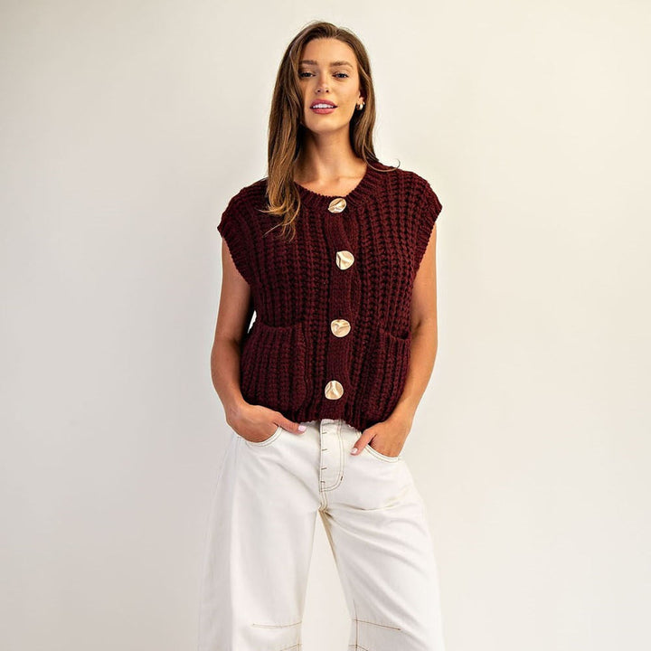 Photograph of a model wearing a chunky burgundy rib-knit vest with large marble-patterned buttons and white wide-leg trousers against a neutral studio backdrop