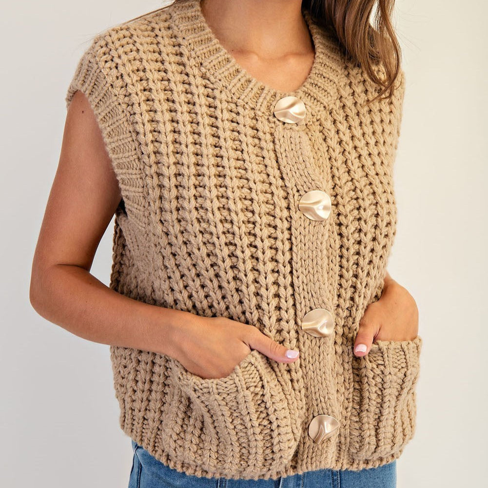 Photograph of a woman wearing a beige chunky knit sweater vest featuring large sculptural wavy buttons and functional front patch pockets against a neutral background