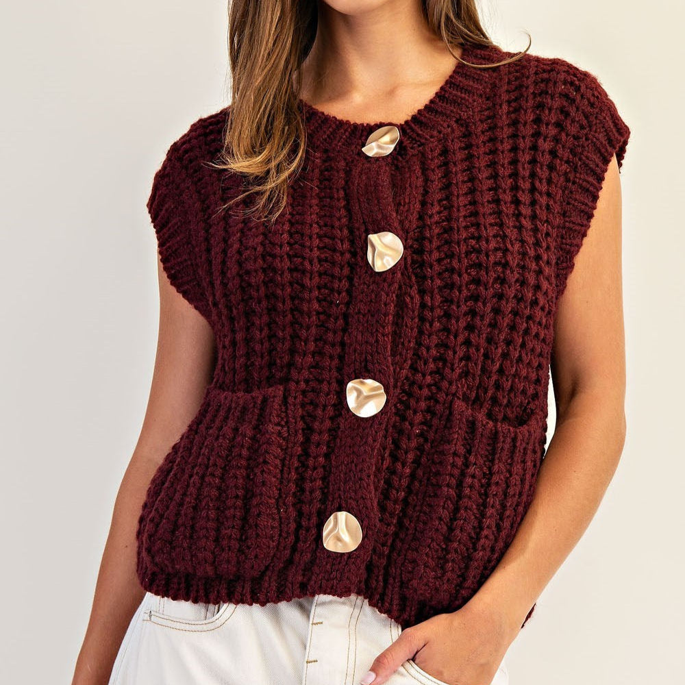 Photograph of a woman in a studio wearing a burgundy chunky knit button-up vest featuring four large irregular gold buttons and front patch pockets paired with white trousers