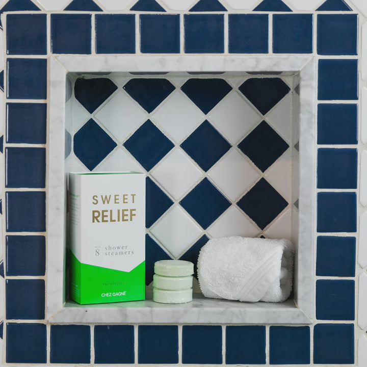 Photograph by Chez Gagné of Sweet Relief eucalyptus shower steamers and a rolled white towel inside a marble-framed shower niche featuring navy blue and white diamond tiles and a stack of three mint green circular discs