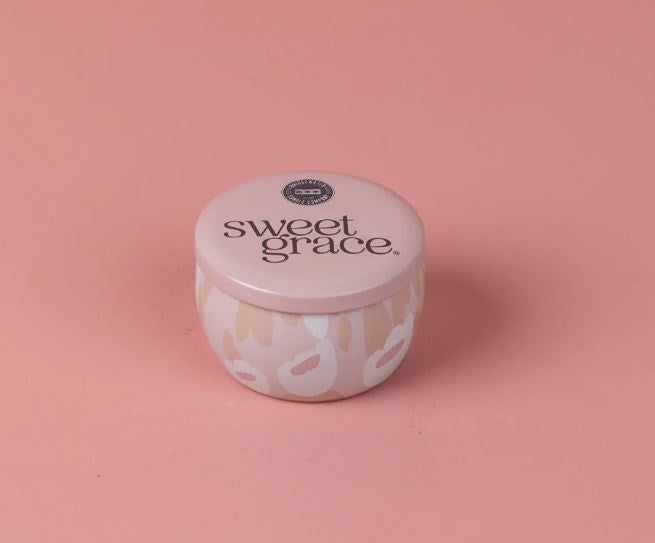 Product photograph of a Bridgewater Candle Company Sweet Grace candle tin on a solid pink background featuring the iconic brand emblem and a decorative abstract petal pattern in muted blush and cream tones