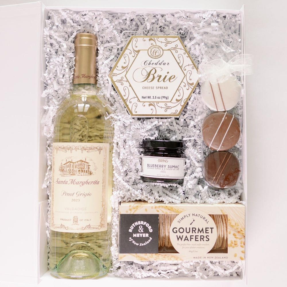 Product photograph of a luxury wine gift hamper in a white box with shredded paper featuring a gold-capped bottle of 2023 Santa Margherita Pinot Grigio, Rutherford & Meyer gourmet wafers, a hexagonal cheddar brie spread, and three chocolate-drizzled treats